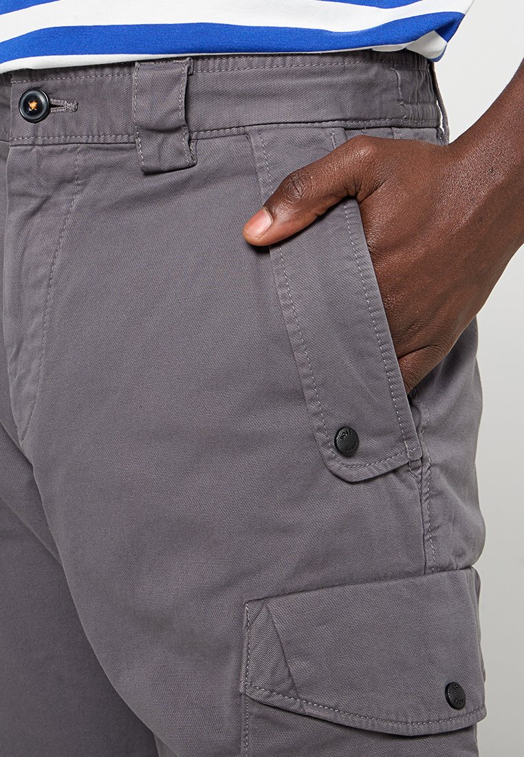 Pantalons cargo gris avec une coupe décontractée, dotés de poches latérales, de boutons métalliques et d'un tissu lisse et texturé. Main placée dans la poche.