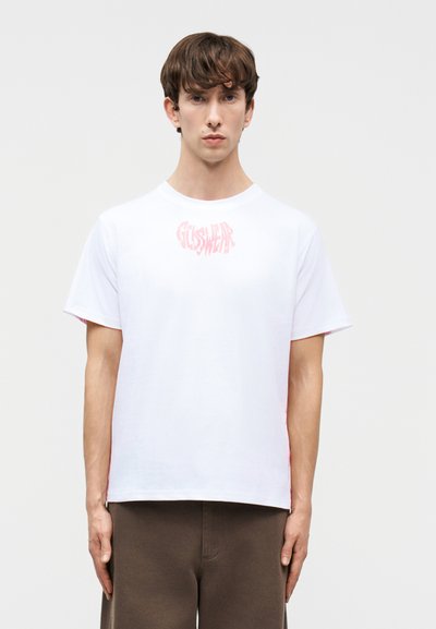 Νέος άνδρας με λευκό κοντομάνικο t-shirt με ροζ λογότυπο "GUSWEAR" και καφέ παντελόνι, στέκεται μπροστά σε απλό φωτεινό φόντο.