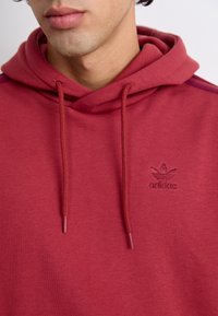 Bordeauxhuva med känguruficka, dragsnöre på huven och en broderad Adidas-logotyp på bröstet. Mjuk textur och avslappnad passform.