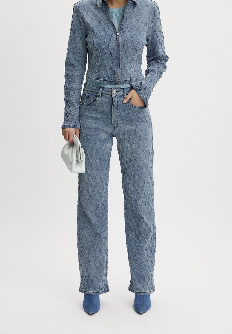 Chaqueta de mezclilla azul acolchada y jeans de tiro alto a juego, con un diseño ajustado y cierre de cremallera. Combinados con un bolso de mano ligero y tacones azules.