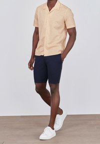 Camicia a maniche corte, a fantasia, di colore arancione chiaro con linee beige, abbinata a pantaloni corti blu scuro e sneakers bianche.