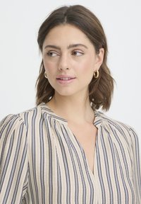 Blouse beige à rayures verticales bleu marine, col en V et épaules plissées. Caractérisée par une texture légère et un design simple.