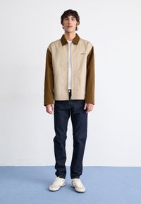 Color-blocked jacket featuring a beige body and brown sleeves, made of sturdy fabric. Paired with dark jeans and light-colored sneakers.
