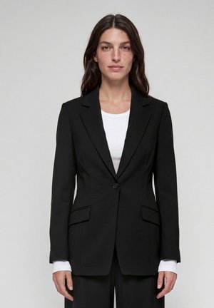 Frau mit langen dunklen Haaren, die einen eng anliegenden schwarzen Blazer über einem weißen Hemd trägt, vor einem schlichten hellen Hintergrund stehend.