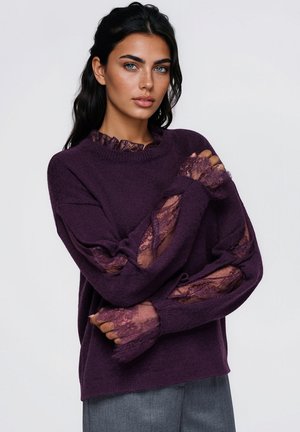 Un mannequin aux longs cheveux foncés pose les bras croisés. Il porte un pull prune à empiècements en dentelle et un pantalon gris, sur un fond clair uni.