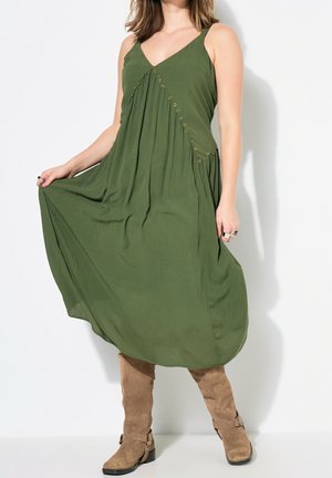 Femme portant une robe midi verte sans manches avec des détails en œillets, tenant l'ourlet, associée à des bottes hauteur genou en suède beige.