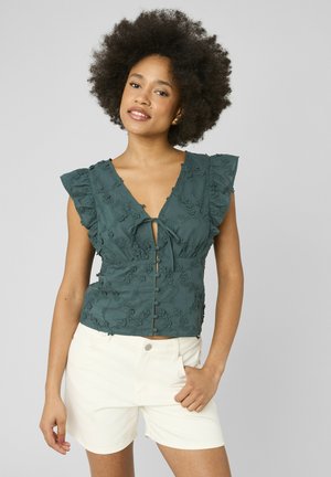 Femme aux cheveux naturellement bouclés portant un chemisier sans manches brodé de fleurs bleu sarcelle et un short blanc, debout devant un fond uni.