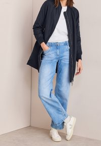 Kobieta w jasnoniebieskich dżinsach, białych trampkach, białej bluzce i długiej czarnej kurtce, stojąca przy beżowej ścianie z jedną ręką w kieszeni.