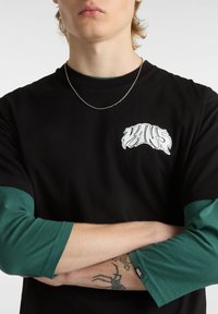 Camiseta negra con el logo "VANS" en blanco, combinada con mangas largas verdes. Características notables incluyen una tela texturizada y un tatuaje visible en el antebrazo.