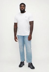 Balti trumpomis rankovėmis marškinėliai su mažu trikampiu logotipu, derinami su šviesiai mėlynais džinsais ir juodais sportbačiais, stovint ant paprasto fono.