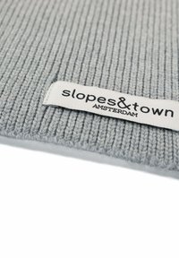 Accessorio in maglia a coste grigia con un'etichetta di stoffa che riporta la scritta "slopes&town AMSTERDAM" in testo nero, caratterizzato da una texture liscia e un design semplice.