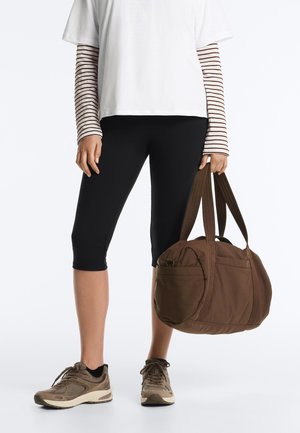 Persona che indossa leggings neri corti, top bianco con maniche lunghe a righe, sneakers beige, che tiene una grande borsa da viaggio marrone per le cinghie.
