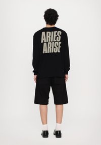 Black long-sleeve shirt with "ARIES ARISE" in bold beige font on the back, paired with black shorts and black shoes with white socks.