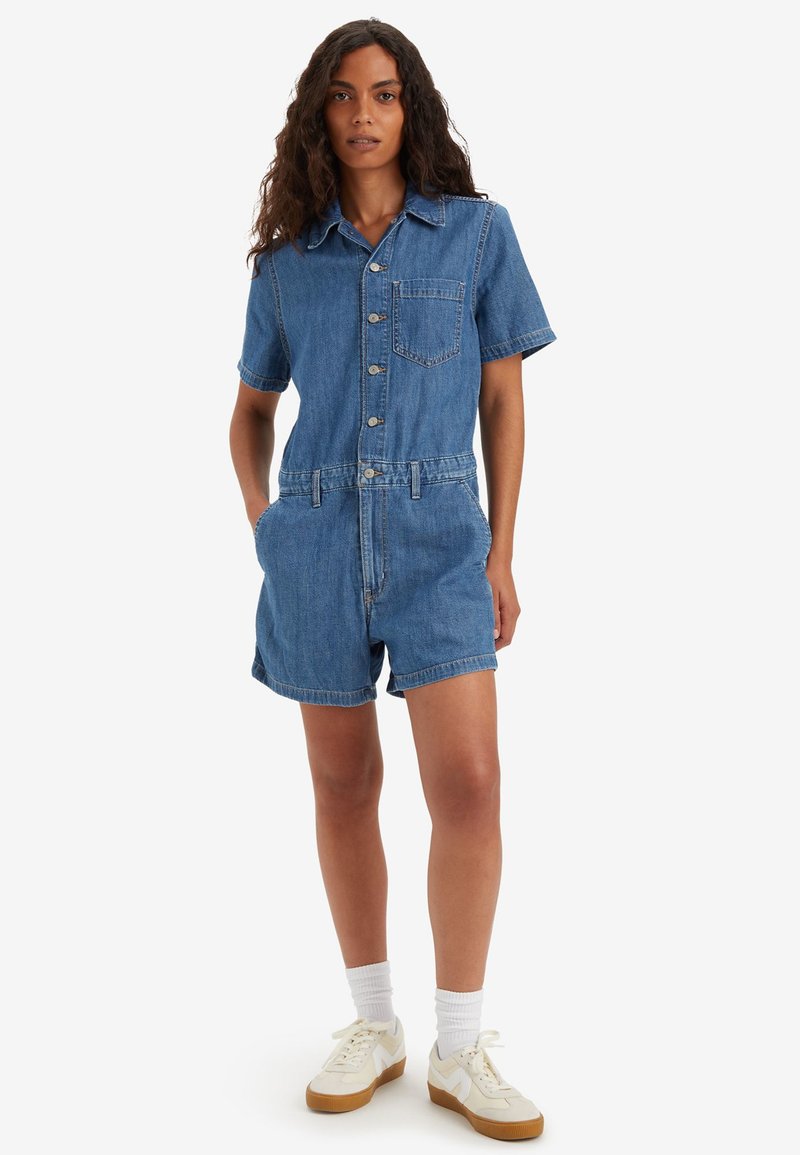 Salopette en denim à manches courtes, bleu clair, avec col, boutons sur le devant et une poche poitrine. Présente une taille cintrée et un short décontracté.