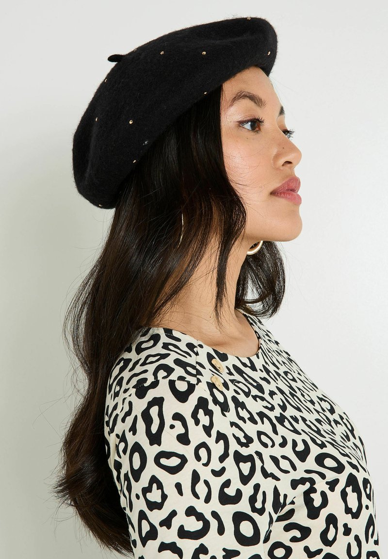 Boina de fieltro negra con tachuelas doradas, de forma redondeada. Se lleva con una blusa crema que presenta un patrón de leopardo negro y acentos de botones.