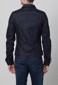 Mörkblå denimjacka med synliga sömmar, figursydd design och knappade ärmslut. Bakifrån syns en smal skärning och fickdetaljer på jeansen.