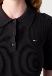 Gros plan d'une personne portant un polo noir côtelé à manches courtes avec trois boutons et un petit logo rouge, blanc et bleu sur la poitrine.