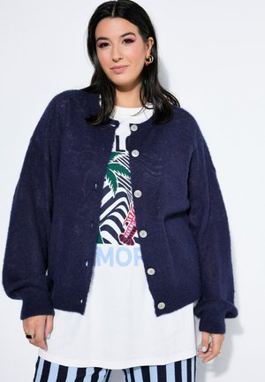 Donna con capelli scuri che indossa un cardigan blu navy abbottonato sopra una T-shirt bianca con grafica e pantaloni a righe, posando davanti a uno sfondo bianco.