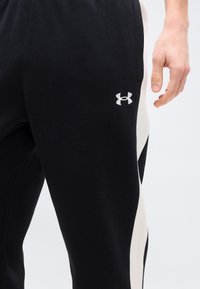 Czarne spodnie sportowe z białymi paskami po bokach i logo Under Armour obok kieszeni, widoczne na prawej nodze i dłoni osoby.