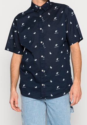 Chemise bleu marine à manches courtes boutonnée, avec motif texte "BEACH CLUB" et chaise de plage blanche, portée avec un jean bleu clair.