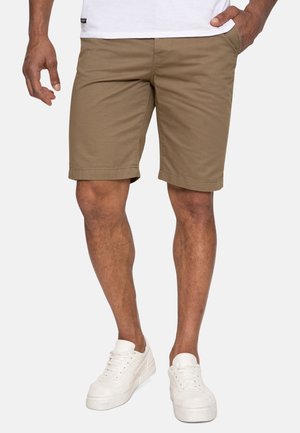 Homme portant un short kaki et des baskets blanches, debout avec une main dans la poche devant un fond uni.