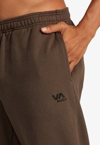 Braune Sweatpants mit elastischem Bund und Seitentasche, mit einem schwarzen, auf dem Oberschenkel gestickten "RVCA"-Logo. Glatte Stofftextur.