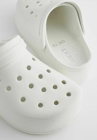 Sabots blancs en caoutchouc texturé, avec des trous de ventilation, un design incurvé et une sangle arrière pour le soutien. Taille UK 12 visible à l'intérieur.