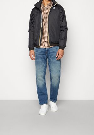 Uomo con giacca nera con zip, camicia marrone a quadri con bottoni, jeans blu e scarpe da ginnastica bianche, in piedi su sfondo bianco.