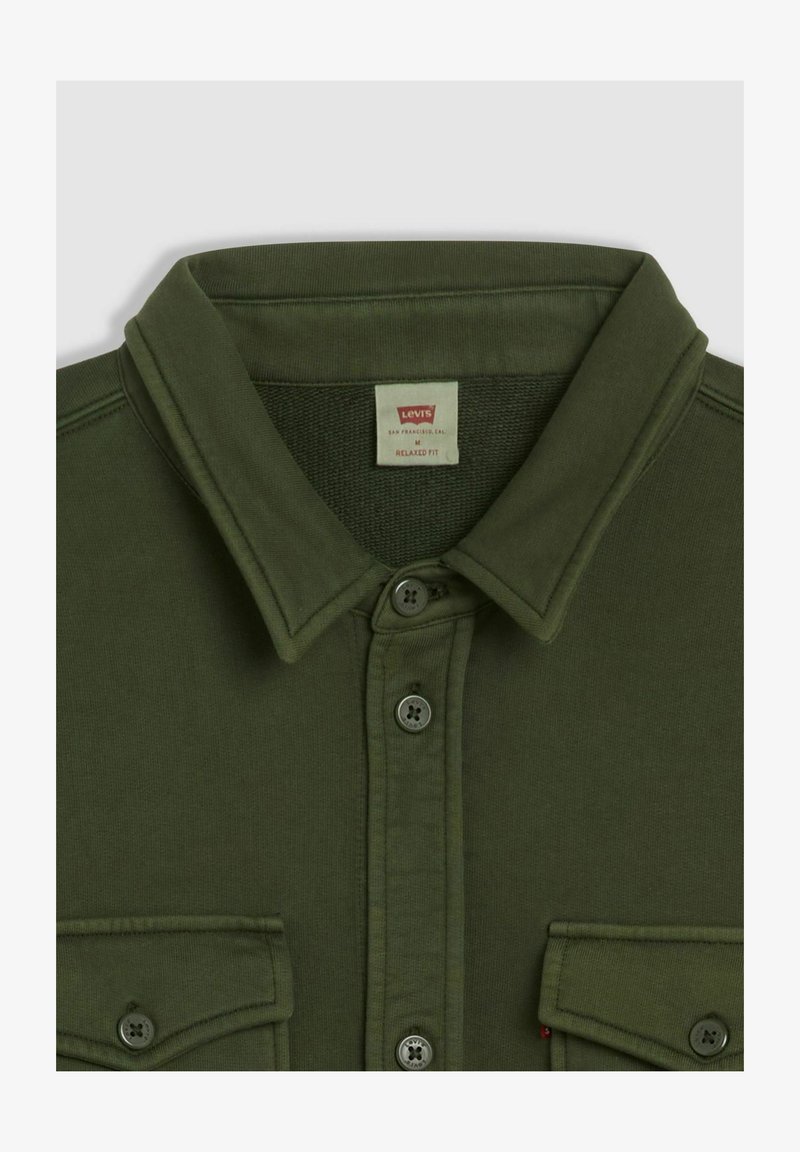 Camicia verde oliva con bottoni, colletto appuntito, due tasche frontali e vestibilità comoda. Tessuto spesso con una texture liscia.