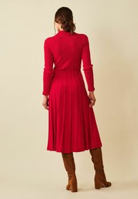 Punainen ribbed-neulottu mekko, jossa on tiukka yläosa ja laskostettu hame, joka ulottuu polveen. Yhdistetty ruskeisiin polven ylle ulottuviin saappaisiin.