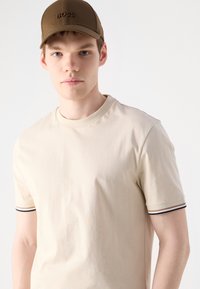 Bežinis medvilninis t-shirt su apykakle ir trumpomis rankovėmis, su kontrastingomis dryžuotomis detalėmis ant rankovių; rudas kepurė su įspaustu logotipu.