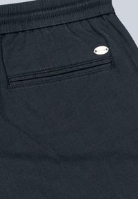 Dunkelblauer Stoff mit einer strukturierten, schrägen Musterung. Verfügt über eine gestickte aufgesetzte Tasche mit einem kleinen silbernen Logo.