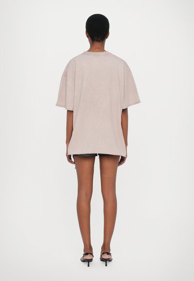 Oversized light beige t-shirt made of soft fabric, featuring a crew neckline and dropped shoulders, paired with dark denim shorts and strappy heels.