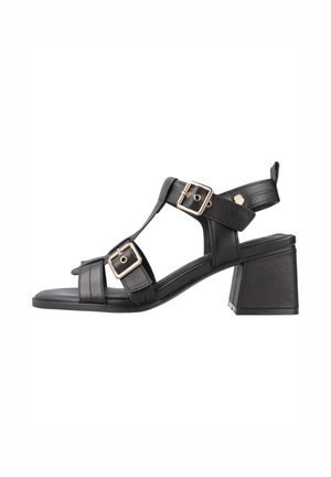 Sandalias de tacón de cuero negro con punta abierta, que presentan dos hebillas ajustables en tono dorado y un diseño de tacón cuadrado. Suela plana, estructura minimalista.