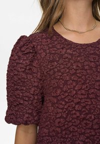 Burgundi textúrájú felső, puffos ujjal és virágmintával, kerek nyakkivágással és sima anyagú felülettel. Dekoratív arany nyaklánc.
