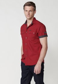 Camisa roja de manga corta hecha de algodón, con un cuello de contraste oscuro, dos bolsillos en el pecho y cierre de botones. Llevada con jeans de mezclilla oscuros.
