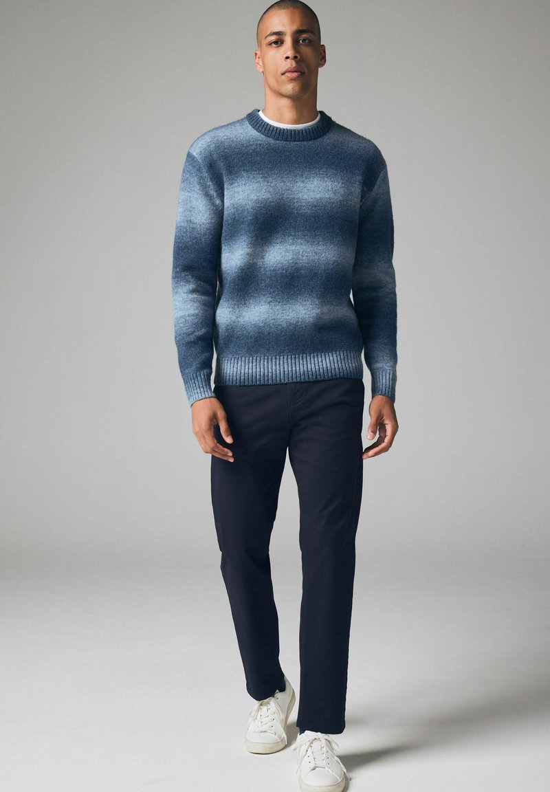 Blau- und grau gestreifter Pullover mit Rundhalsausschnitt und gerippten Bündchen, kombiniert mit dunklen Hosen und weißen Sneakers.