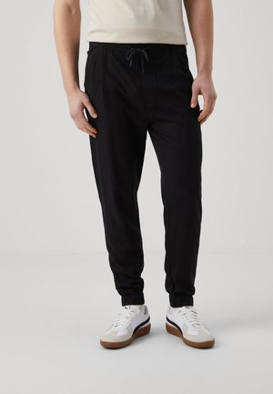 Man draagt zwarte joggingbroek met elastische boorden, witte sneakers met bruine zolen, staand tegen een effen lichte achtergrond.