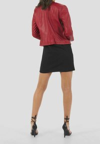 Veste en cuir rouge à coupe ajustée, associée à une mini-jupe noire. Le mannequin porte des sandales à talons hauts noires avec des lacets autour de la cheville.