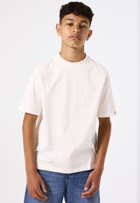 Camiseta de algodón crema con cuello redondo y mangas cortas, que presenta un parche circular azul en el lado derecho, combinada con jeans azules.