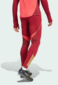Rote Sportleggings mit gelben Akzenten, die eine schmal zulaufende Passform und Reißverschlussbündchen haben. Kombiniert mit schwarzen Schuhen und weißen Socken.