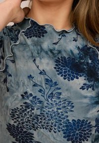 Blomstret bluse i blå og grå med en flæset halsudskæring. Har indviklede mørkeblå blomster mønstre på et blødt, tekstureret stof.