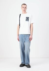 Camiseta blanca con un bolsillo de patrón en blanco y negro, combinada con unos vaqueros holgados de color azul claro. La figura lleva una bandolera negra y zapatos oscuros.