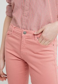 Jeans rosa claro de denim, con un diseño ajustado, cierre de botón metálico y bolsillos frontales. Camisa rayada parcialmente visible.