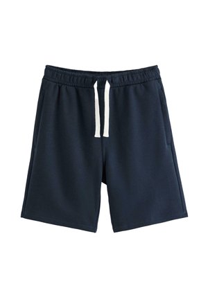 Chłopiec ubrany w beżową czapkę z daszkiem założoną do tyłu, białą koszulkę polo, granatowe spodenki, białe skarpetki i beżowe buty sportowe, stojący i patrzący na bok.