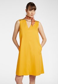 Abito giallo senza maniche con scollatura a V, caratterizzato da bottoni decorativi sulle spalle e due tasche laterali. Accompagnato da una sciarpa a fantasia.