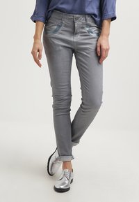 Šedé džíny s úzkým střihem, zdobené kovově modrými akcenty na kapsách a mírným leskem. V kombinaci se stříbrnými šněrovacími botami.