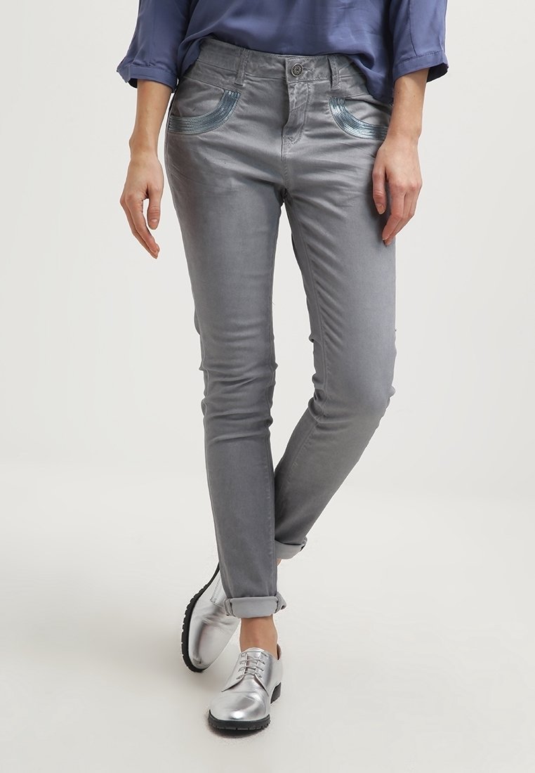 Šedé džíny s úzkým střihem, zdobené kovově modrými akcenty na kapsách a mírným leskem. V kombinaci se stříbrnými šněrovacími botami.