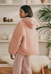 Rosa flauschige Kapuzenjacke aus strukturiertem Stoff, im Oversized-Schnitt, mit kleinen Ohren an der Kapuze. Kombiniert mit gestreiften Hosen in Pink und Weiß.