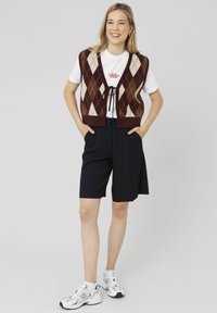 Gilet en tricot brun à motif argyle avec des losanges blancs et beiges, T-shirt blanc en dessous, associé à un short marine à rayures et des baskets blanches.